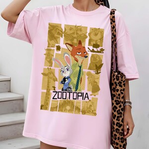 May include: A light pink t-shirt featuring a graphic from the Disney movie Zootopia. The design includes characters Judy Hopps and Nick Wilde, with the movie title in bold letters. The shirt has short sleeves and a relaxed fit.