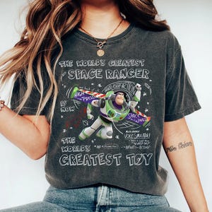 Op de afbeelding: Donkergrijze t-shirt met een afbeelding van Buzz Lightyear en de tekst "THE WORLD'S GREATEST SPACE RANGER" en "THE WORLD'S GREATEST TOY". Het ontwerp bevat de zinnen "HIGH PRESSURE" en "SPACE WINGS".