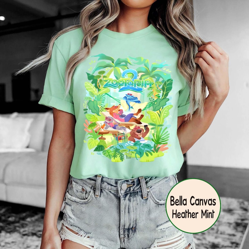 Pode incluir: Camiseta verde claro com um gr&aacute;fico colorido de "Zootopia". O design apresenta personagens e plantas tropicais. A camiseta &eacute; feita de um material macio e &eacute; rotulada como "Bella Canvas Heather Mint".