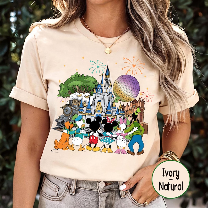 Peut inclure: T-shirt de couleur ivoire avec un motif color&eacute; de personnages et de monuments Disney, dont un ch&acirc;teau, un train et Spaceship Earth. Le motif comprend Mickey et Minnie Mouse, Donald Duck et Dingo. Le t-shirt a des manches courtes et un col rond.
