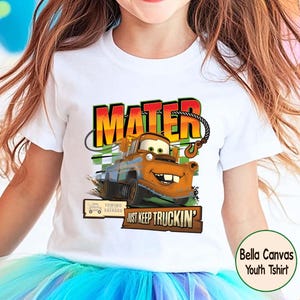 Puede incluir: Camiseta blanca Bella Canvas para j&oacute;venes con un gr&aacute;fico de Mate de la pel&iacute;cula Cars. El dise&ntilde;o incluye el nombre "MATER" en naranja y rojo, y la frase "JUST KEEP TRUCKIN'". La camiseta tiene cuello redondo.