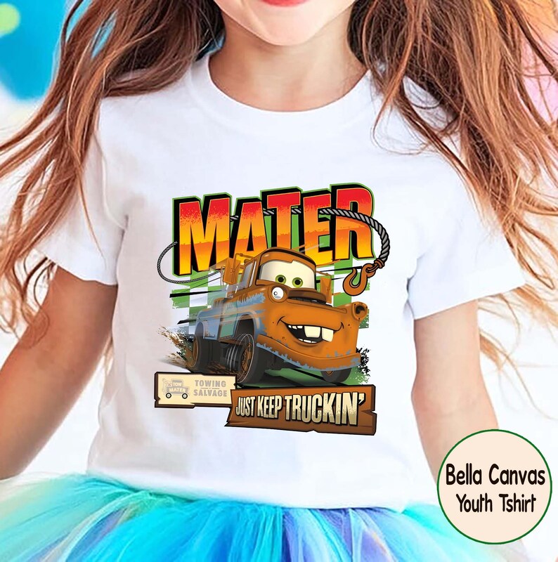 Puede incluir: Camiseta blanca Bella Canvas para j&oacute;venes con un gr&aacute;fico de Mate de la pel&iacute;cula Cars. El dise&ntilde;o incluye el nombre "MATER" en naranja y rojo, y la frase "JUST KEEP TRUCKIN'". La camiseta tiene cuello redondo.