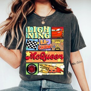 Puede incluir: Camiseta gris oscuro con un dise&ntilde;o gr&aacute;fico colorido. El dise&ntilde;o incluye las palabras "Lightning Ka-Chow! McQueen" con im&aacute;genes de un coche de carreras, una bandera a cuadros, un trofeo y el n&uacute;mero 95 con llamas. La camiseta tambi&eacute;n tiene un logotipo de Lightyear.