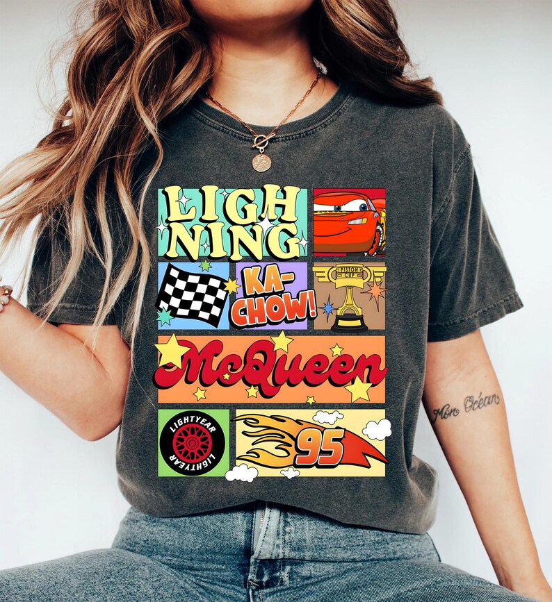 Puede incluir: Camiseta gris oscuro con un dise&ntilde;o gr&aacute;fico colorido. El dise&ntilde;o incluye las palabras "Lightning Ka-Chow! McQueen" con im&aacute;genes de un coche de carreras, una bandera a cuadros, un trofeo y el n&uacute;mero 95 con llamas. La camiseta tambi&eacute;n tiene un logotipo de Lightyear.