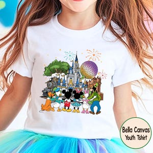 Peut inclure: T-shirt blanc pour enfant avec un graphisme color&eacute; de personnages Disney devant un ch&acirc;teau, un train et une sph&egrave;re. Le motif comprend des feux d'artifice et le texte "Bella Canvas Youth Tshirt".