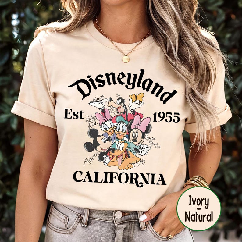 May include: Ivory-colored t-shirt featuring the Disneyland logo with the text "Est 1955 CALIFORNIA". The shirt showcases a graphic of Disney characters including Mickey Mouse, Minnie Mouse, Donald Duck, and Goofy. The shirt is ivory natural.