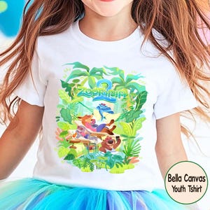 Pode incluir: Camiseta branca juvenil com um gr&aacute;fico colorido de Zootopia. O design apresenta personagens como Nick Wilde e Judy Hopps, cercados por folhagens tropicais. O texto "Zootopia 2" tamb&eacute;m &eacute; vis&iacute;vel. A camiseta &eacute; rotulada como "Bella Canvas Youth Tshirt".