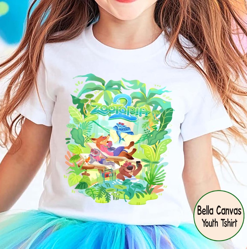 Pode incluir: Camiseta branca juvenil com um gr&aacute;fico colorido de Zootopia. O design apresenta personagens como Nick Wilde e Judy Hopps, cercados por folhagens tropicais. O texto "Zootopia 2" tamb&eacute;m &eacute; vis&iacute;vel. A camiseta &eacute; rotulada como "Bella Canvas Youth Tshirt".