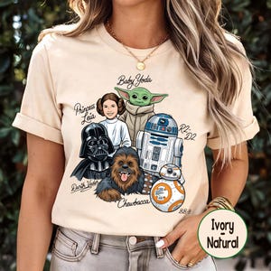 May include: Ivory-colored t-shirt featuring a graphic of Star Wars characters: Baby Yoda, Princess Leia, Darth Vader, Chewbacca, R2-D2, and BB-8. The shirt has short sleeves and a round neckline. The words "Ivory Natural" are printed on a circular label.