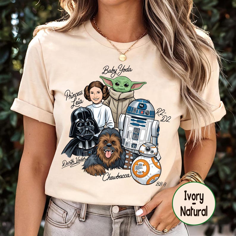May include: Ivory-colored t-shirt featuring a graphic of Star Wars characters: Baby Yoda, Princess Leia, Darth Vader, Chewbacca, R2-D2, and BB-8. The shirt has short sleeves and a round neckline. The words "Ivory Natural" are printed on a circular label.