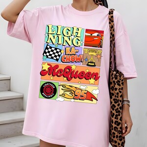 Puede incluir: Camiseta rosa claro con un dise&ntilde;o gr&aacute;fico colorido. El dise&ntilde;o incluye el texto "LIGHTNING KA-CHOW! McQueen" junto con im&aacute;genes de un coche de carreras, una bandera a cuadros, un trofeo, un neum&aacute;tico y el n&uacute;mero 95. La camiseta es de corte holgado.