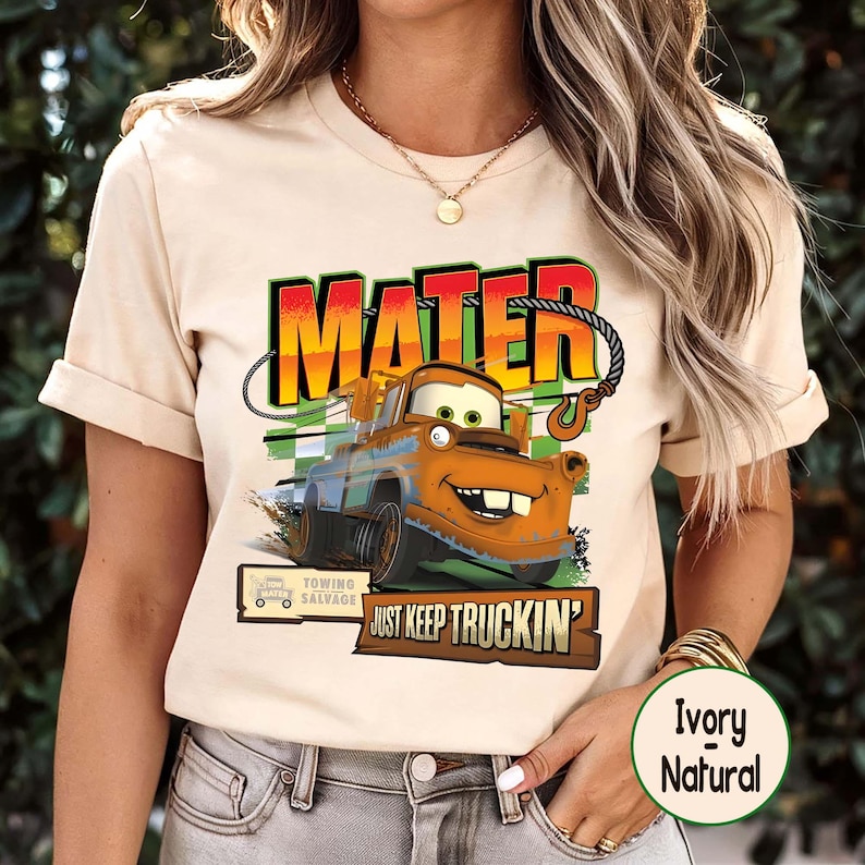 Puede incluir: Camiseta de color marfil con un dise&ntilde;o de Mate de la pel&iacute;cula Cars. El dise&ntilde;o incluye la palabra "MATER" en negrita, una ilustraci&oacute;n de un cami&oacute;n de remolque y la frase "JUST KEEP TRUCKIN'". La camiseta tambi&eacute;n tiene un logotipo de "Towing Salvage".
