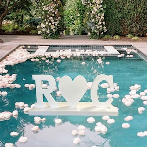 May include: A white floating sign in a pool with the letters "R" and "C" flanking a heart shape. The pool is surrounded by white flower petals and a floral archway. The water is a bright turquoise color.