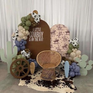 May include: A decorative event display featuring a brown arched backdrop with the text A LITTLE COWBOY IS ON THE WAY, a patterned arch, a wicker chair, a cowhide rug, balloon garlands in olive, blue, and cow print, and two light green cactus cutouts.