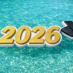 May include: A golden glitter-covered "2026" floats in turquoise water, accompanied by a black graduation cap with a gold tassel. The numbers are outlined in white, creating a 3D effect. The image evokes a graduation theme.