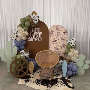 May include: A decorative event display featuring a rattan chair, a cowhide rug, and two arched backdrops. One brown arch displays the text A LITTLE COWBOY IS ON THE WAY. The scene includes balloon clusters in olive, tan, blue, and cow-print patterns.