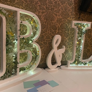 May include: White wooden letters spelling "B & L" decorated with greenery and lights. The letters are on a white table with a few cards on the surface.