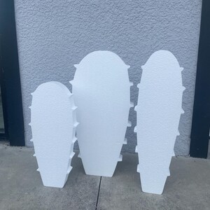 May include: Three white, textured, foam cactus-shaped cutouts of varying heights. Each piece features a rounded top, a tapered base, and small, protruding, triangular-shaped spikes along the sides, creating a uniform, matte, and lightweight appearance.