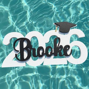 May include: A white wooden graduation decoration with the year "2025" and the name "Brooke" in black script. A small black graduation cap with a gold tassel sits atop the "5". The decoration floats on a turquoise water surface.
