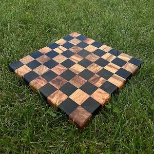 May include: A black and brown wooden chessboard with a checkered pattern. The board is made of wood with a natural grain and is sitting on a green grassy surface.