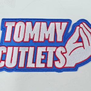 May include: Embroidered patch with the words "TOMMY CUTLETS" in white, outlined in red, on a blue background. The patch features a white hand gesture, outlined in red, with the fingers and thumb touching. The patch has a blue border.