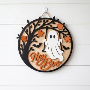 Op de afbeelding: Een rond houten wandbord met een zwart silhouet van een boom, oranje esdoornbladeren en twee zwarte vleermuizen. Een wit spookje zit op een schommel aan de takken. De tekst Hey Boo staat in oranje letters onderaan het bord afgebeeld.