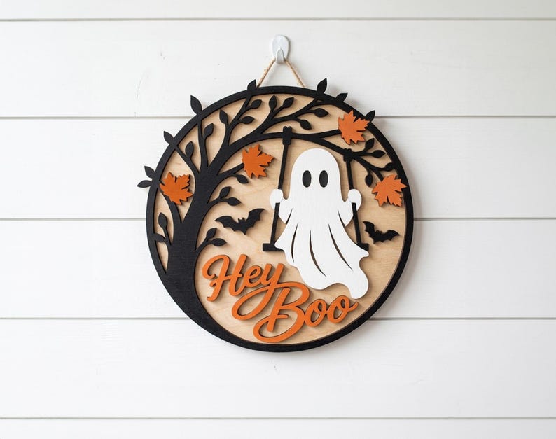 Op de afbeelding: Een rond houten wandbord met een zwart silhouet van een boom, oranje esdoornbladeren en twee zwarte vleermuizen. Een wit spookje zit op een schommel aan de takken. De tekst Hey Boo staat in oranje letters onderaan het bord afgebeeld.