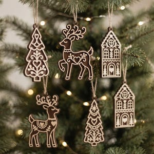May include: A set of six dark brown wooden ornaments featuring intricate white laser-cut designs of reindeer, pine trees, and houses. Each ornament is suspended by a thin jute twine loop, showcasing detailed folk-inspired patterns and heart motifs.