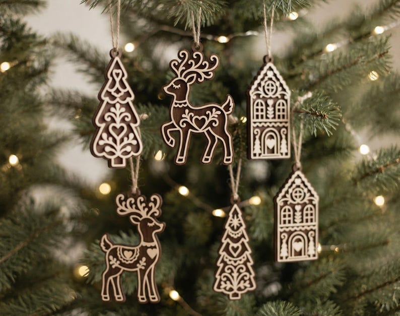 May include: A set of six dark brown wooden ornaments featuring intricate white laser-cut designs of reindeer, pine trees, and houses. Each ornament is suspended by a thin jute twine loop, showcasing detailed folk-inspired patterns and heart motifs.