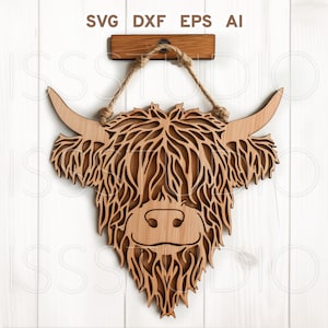 Puede incluir: Un recorte de madera de la cabeza de una vaca Highland con un colgador de cuerda. La vaca tiene un patrón de pelaje marrón y blanco y una expresión amigable.