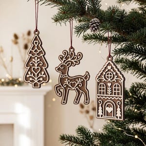 May include: Three dark brown wooden ornaments hanging from a tree. The ornaments feature intricate laser-cut designs including a stylised Christmas tree, a reindeer with large antlers, and a house with windows and a door, all suspended by thin red strings.