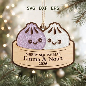 Pode incluir: Um ornamento de madeira para pendurar com dois personagens de bolinhos brilhantes e sorridentes numa cesta de vapor. A base de madeira clara está gravada com o texto MERRY SQUISHMAS, os nomes Emma & Noah e o ano 2026. O ornamento pende de um fio.