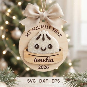 Może przedstawiać: Okrągła drewniana ozdoba świąteczna z brokatową białą pierożkową postacią w koszyku do gotowania na parze. Napis głosi MY SQUISHY YEAR, Amelia oraz 2026. Beżowa kokarda z tkaniny jest przymocowana u góry za pomocą sznurka do zawieszenia.