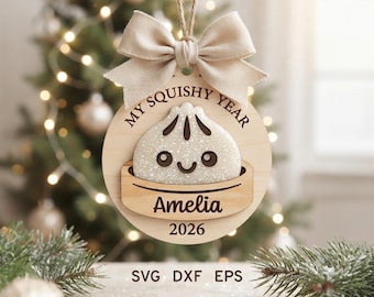 Könnte beinhalten: Ein runder Weihnachtsanhänger aus Holz mit einem glitzernden weißen Knödel-Charakter in einem Dampfkorb. Der Text lautet MY SQUISHY YEAR, Amelia und 2026. Eine beigefarbene Stoffschleife ist oben mit einer Kordel zum Aufhängen befestigt.
