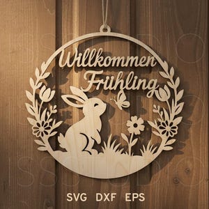 Może przedstawiać: Okrągły drewniany znak z niemieckim napisem "Willkommen Frühling", królikiem, motylem i kwiatowym wzorem. Znak wykonany jest z jasnego drewna i przeznaczony do zawieszenia.