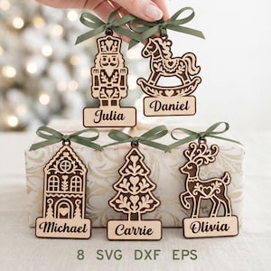 Puede incluir: Cinco adornos navideños de madera cortados con láser que representan un cascanueces, un caballito, una casa, un árbol y un reno. Cada adorno tiene un lazo de cinta verde y una base rectangular con los nombres Julia, Daniel, Michael, Carrie y Olivia.