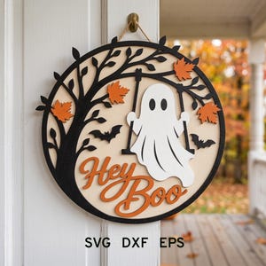 May include: A circular wooden wall sign featuring a black tree silhouette, orange maple leaves, two black bats, and a white ghost on a swing. The text "Hey Boo" is written in orange script. The sign includes the text SVG DXF EPS at the bottom.