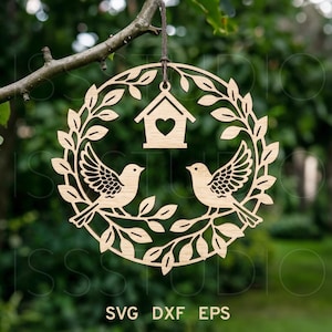 Puede incluir: Decoración de madera circular con temática de aves. El diseño presenta dos pájaros posados en ramas dentro de una corona de hojas. Una pequeña casa para pájaros con un recorte en forma de corazón está en la parte superior. El texto "SVG DXF EPS" está en la parte inferior.