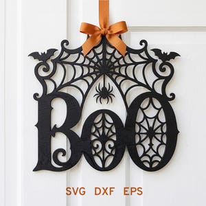 May include: A black laser-cut wall hanging featuring the word BOO with intricate spider web patterns, two small bats at the top corners, and a central spider. The piece is suspended by an orange ribbon bow. Text at the bottom reads SVG DXF EPS.