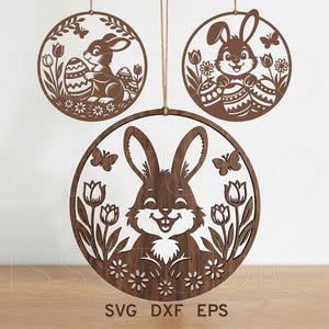 May include: Three wooden Easter decorations featuring bunny rabbits, eggs, flowers, and butterflies. The designs are cut out of brown wood and hang from twine. The bottom decoration has the text "SVG DXF EPS".