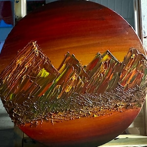 May include: A circular painting depicting a mountain range at sunset. The mountains are painted in shades of brown, green, and yellow, with a textured, brushstroke effect. The sky is a vibrant orange and red, creating a warm and inviting atmosphere.