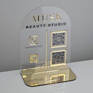 Op de afbeelding: Een helder, boogvormig acrylbord met een goudkleurige spiegelvoet. Het bord bevat de tekst MUSE BEAUTY STUDIO, een Instagram-pictogram met de handle mmariana_mua, een goudkleurig vierkant logo en twee QR-codes met een gouden omlijsting.