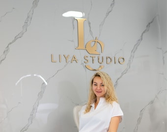 May include: A person with blonde hair wearing a white short-sleeved collared shirt and white trousers, sitting in a white chair against a light gray marble wall featuring the gold-colored logo and text LIYA STUDIO in a metallic finish.