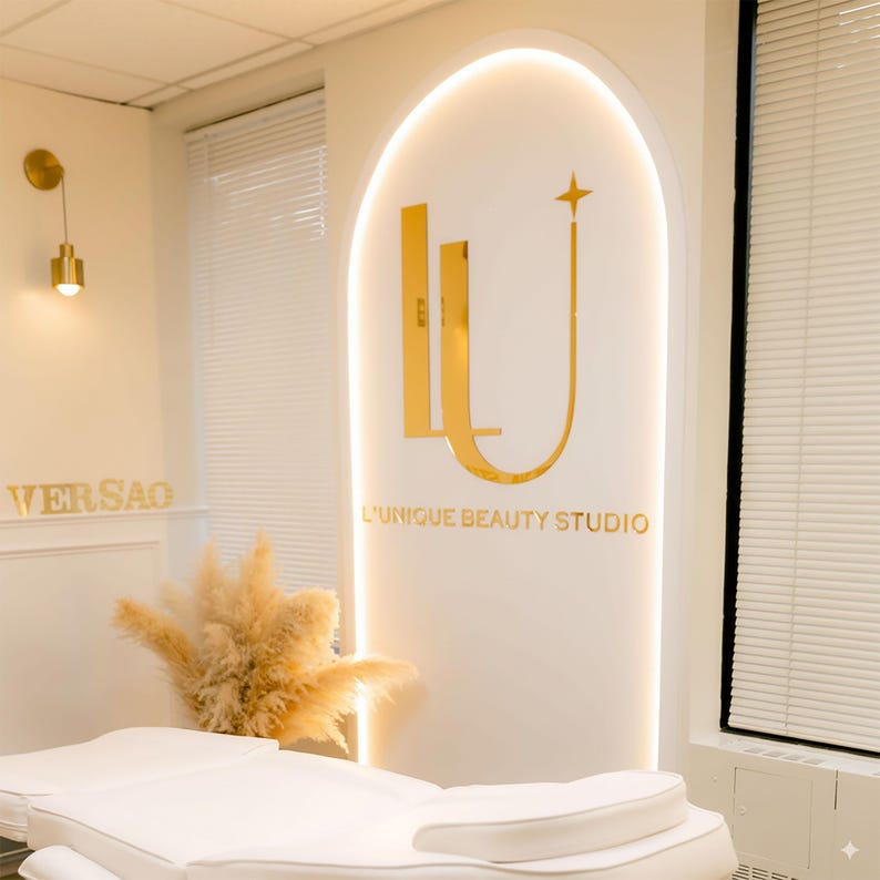 Może przedstawiać: Wnętrze studia urody z białym ł&oacute;żkiem zabiegowym. Na ścianie złote logo z napisem "L'Unique Beauty Studio". Dekoracyjne oświetlenie i trawa pampasowa dopełniają estetykę. Dominująca kolorystyka to biel i złoto.