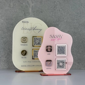 Può includere: Due insegne in acrilico personalizzate con codici QR. La più grande, color crema a forma di arco, mostra "Hair By Emma J Chaney" con icone Instagram e TikTok. La più piccola, rosa e di forma organica, mostra "SASSY med spa" con icone Zelle e Venmo.