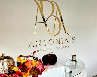 May include: A gold sign with the text "ANTONIA'S BEAUTY ACADEMY" above a stylized gold "AB" logo. A white counter with a glass top is decorated with fall foliage and a burgundy pumpkin.