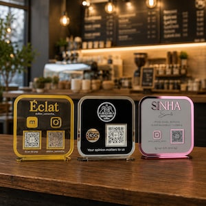 Puede incluir: Tres letreros cuadrados de acrílico con esquinas redondeadas y soportes. El letrero izquierdo es dorado con el texto Éclat, iconos de redes sociales y dos códigos QR. El letrero central es negro con un logotipo, un código QR y el texto Your opinion matters to us. El letrero derecho es rosa con el texto SINHA Events, iconos de redes sociales, un código QR y un número de teléfono.