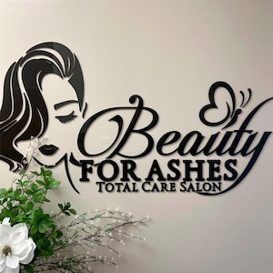 Może przedstawiać: Czarny akrylowy szyld salonu z napisem "Beauty For Ashes Total Care Salon" w stylowej czcionce. Znak przedstawia sylwetkę kobiety i motyw motyla. Zielono-biała kompozycja kwiatowa na pierwszym planie.