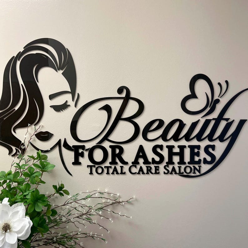 Może przedstawiać: Czarny akrylowy szyld salonu z napisem "Beauty For Ashes Total Care Salon" w stylowej czcionce. Znak przedstawia sylwetkę kobiety i motyw motyla. Zielono-biała kompozycja kwiatowa na pierwszym planie.