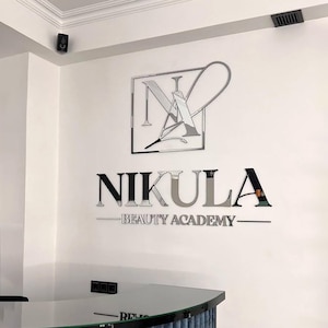 Może przedstawiać: Biała ściana z logo "Nikula Beauty Academy". Logo zawiera stylizowane "N" i "A" w kwadratowej ramie, z nazwą akademii i hasłem w srebrnych i czarnych literach.